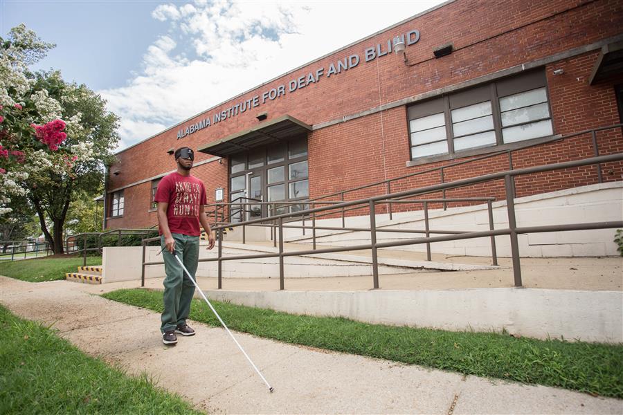 A student using a white cane to walk in front of an A.I.D.B. building.